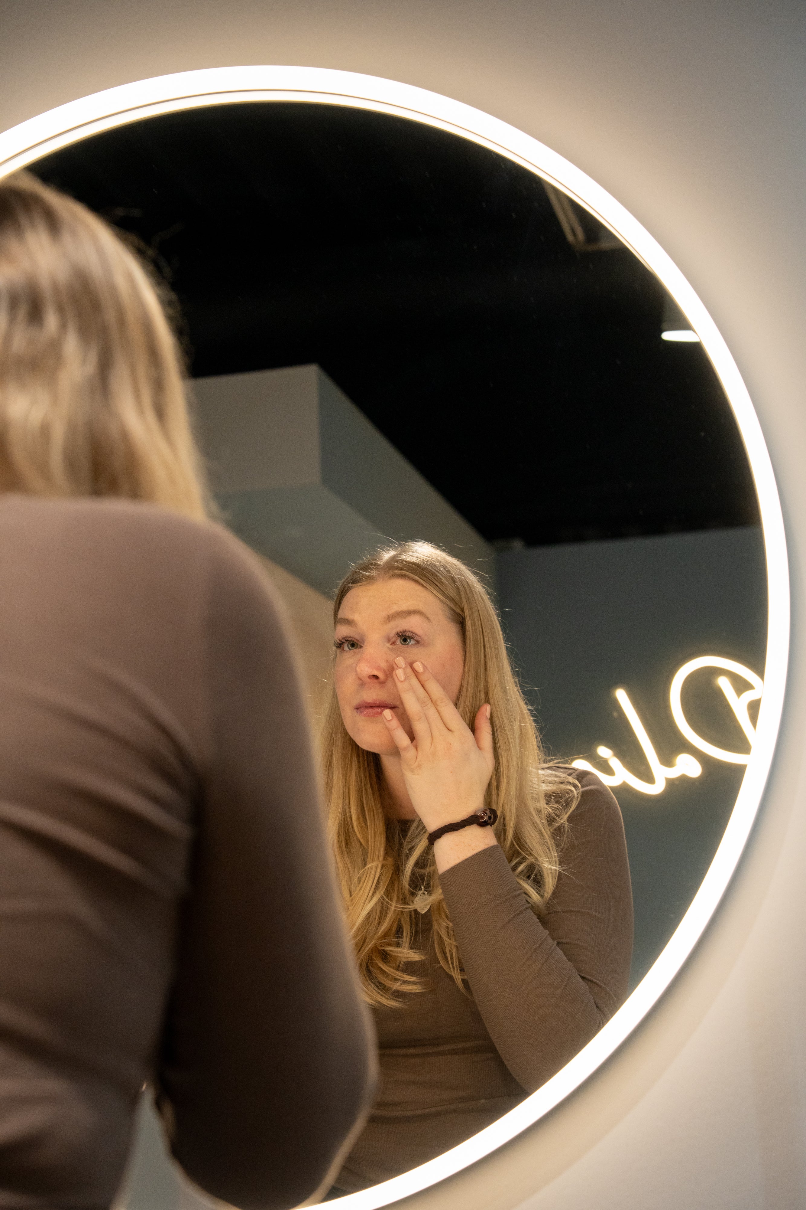 Frau steht vor einem Spiegel und trägt ein Produkt in ihrem Gesicht auf. 